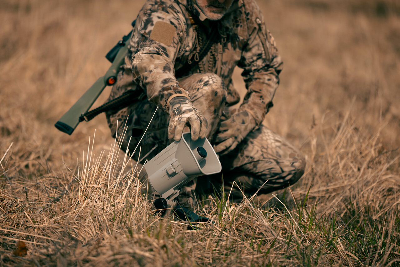 Electronic predator call unit placed on open ground at dusk with a coyote hunting setup in the background